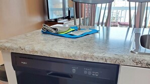 Cocina compacta con lavavajillas integrado, una mesita con papeles y un televisor sobre un mueble.