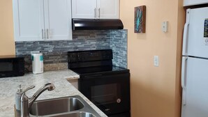 Una cocina con armarios blancos, horno negro, pileta de acero inoxidable y refrigerador blanco.