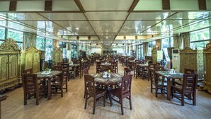 Uma ampla sala de jantar com várias mesas e cadeiras, grandes janelas e mobiliário de madeira ornamentado.