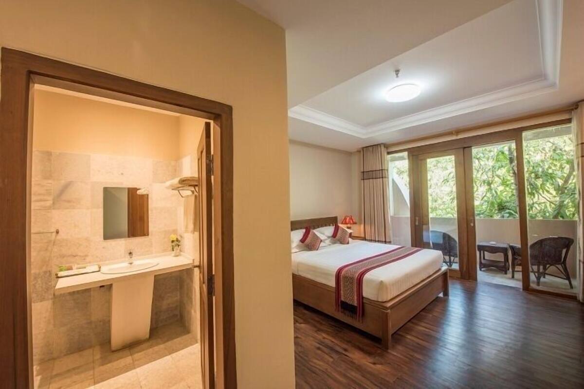 Een hotelkamer met een houten bed, een badkamer met een wastafel en spiegel, en een balkon met uitzicht op groen.