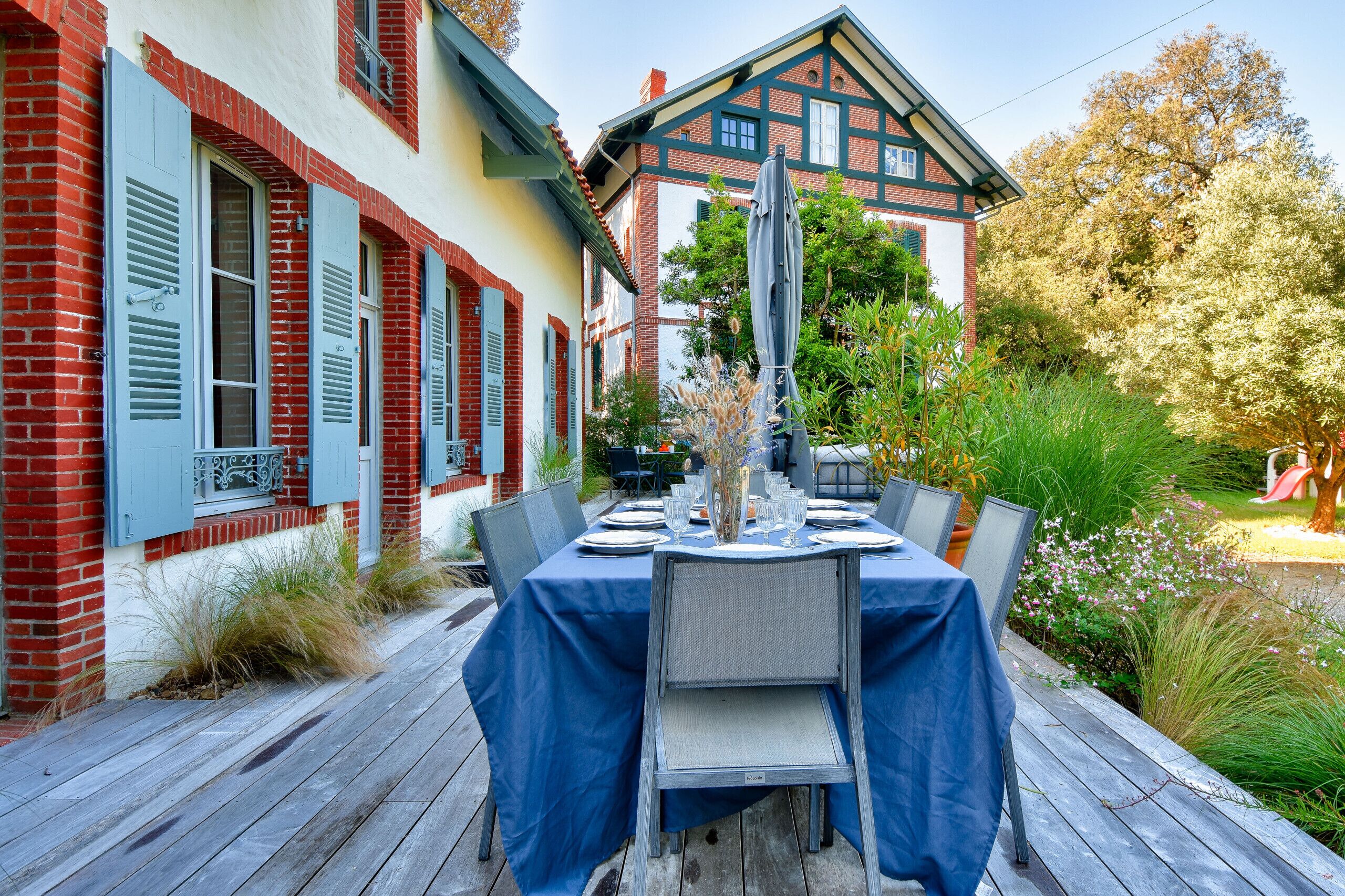 An outdoor dining area with a table set for six, surrounded by chairs, in front of a building with red brick and white walls.