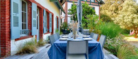 An outdoor dining area with a table set for six, surrounded by chairs, in front of a building with red brick and white walls.