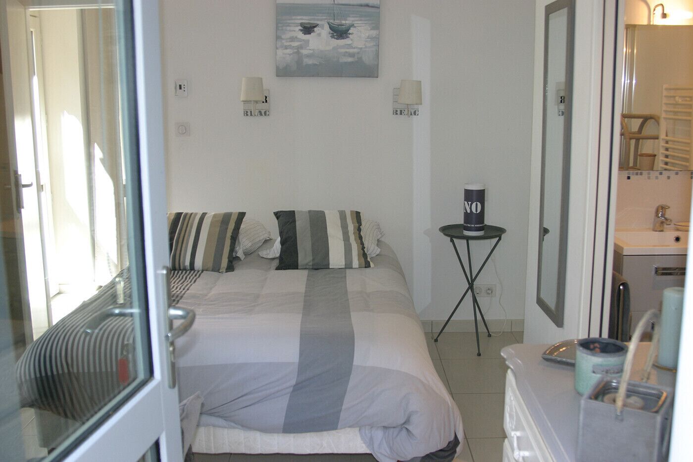 A bedroom with a bed, pillows, a bedside table with a lamp, a painting on the wall, and a bathroom visible through an open door.