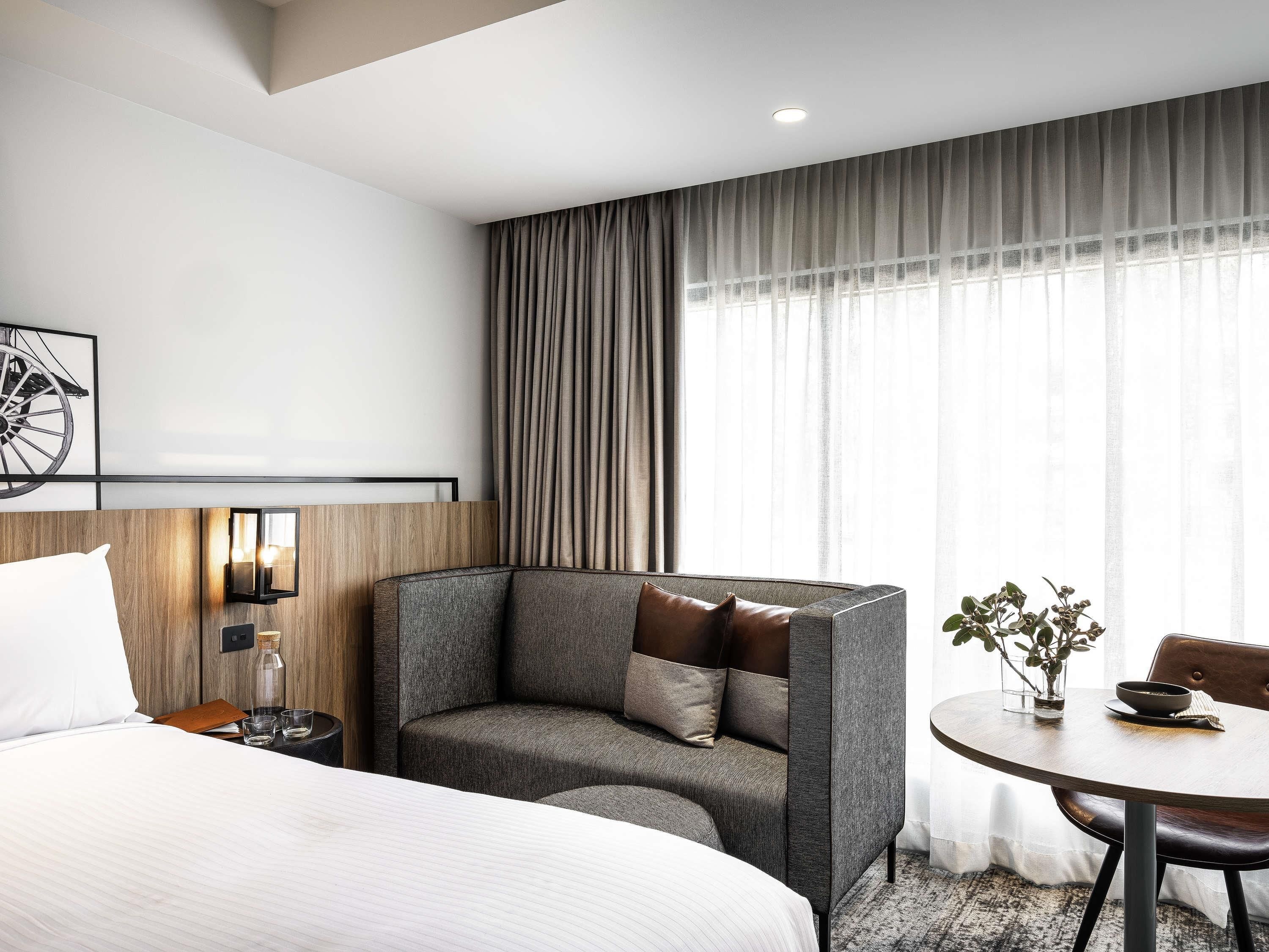 A hotel room with a bed, a sofa, a dining table, and a large window with curtains.
