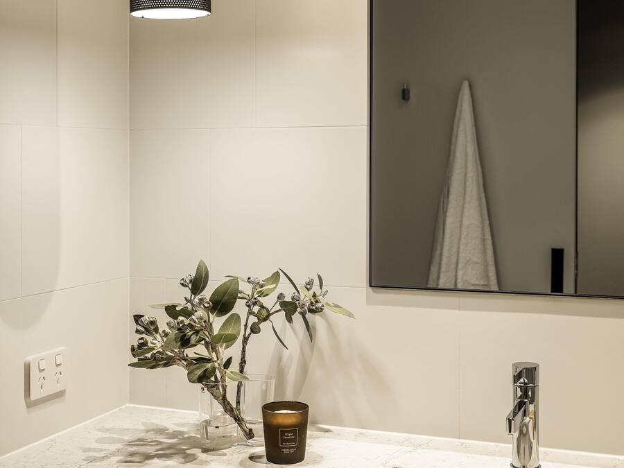 Un baño con pared de azulejos blancos, un espejo, una planta, una vela y un grifo.