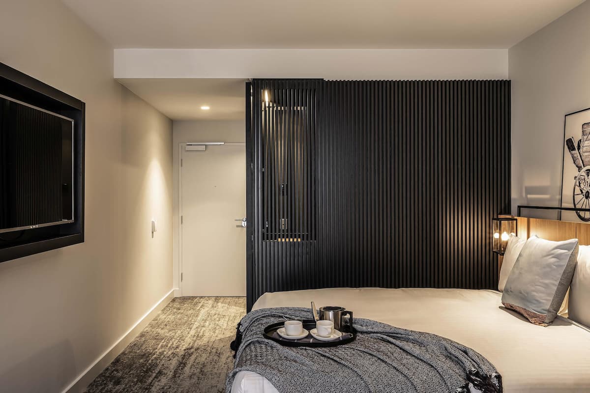 A modern hotel room with a bed, a tray with cups and a kettle, a framed picture on the wall, and a door with a vertical panel design.