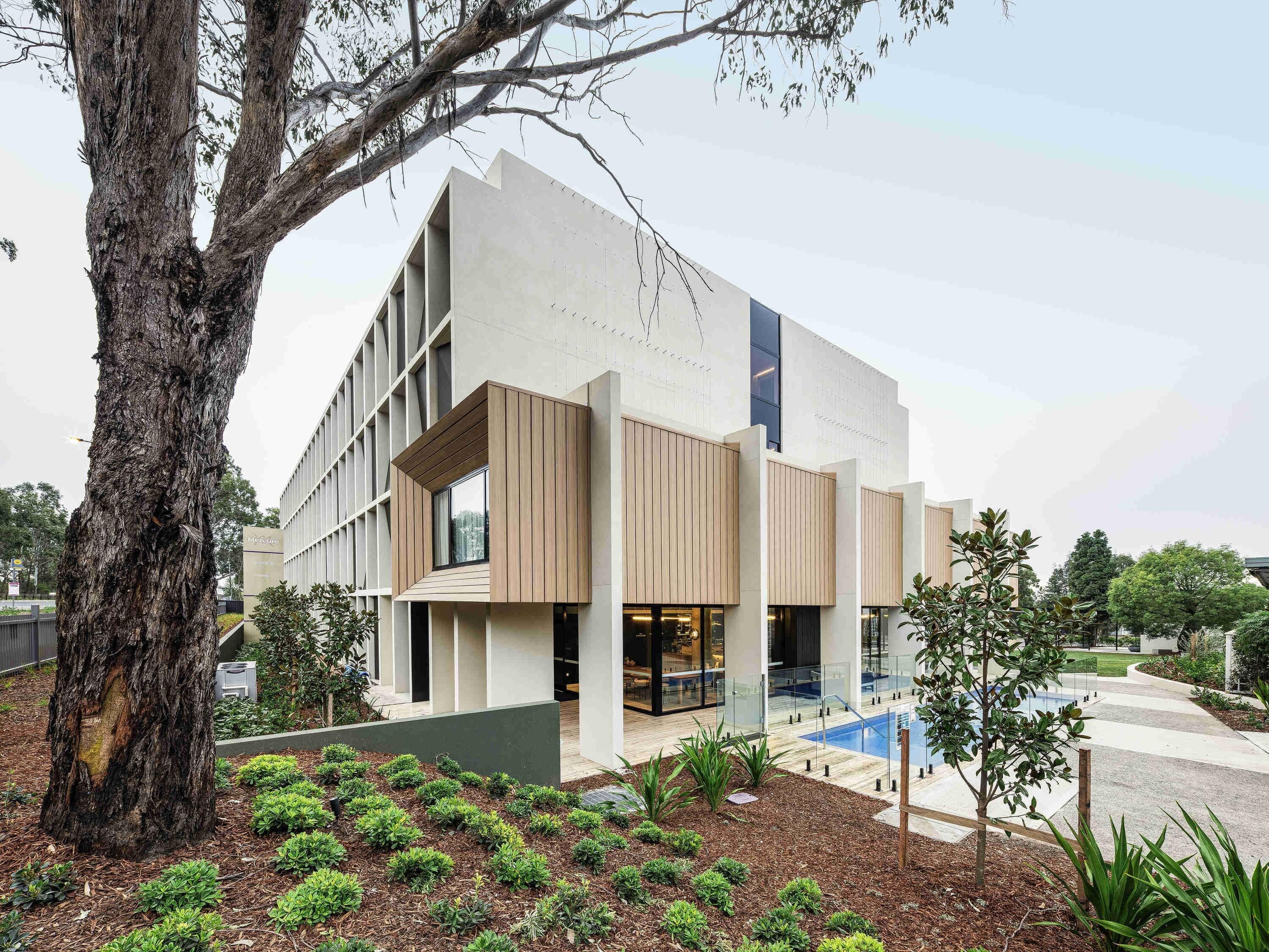 Un edificio moderno con un gran árbol en primer plano, un jardín ajardinado y un cielo despejado.