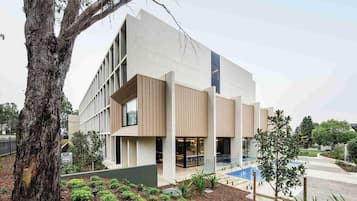 Un edificio moderno con un gran árbol en primer plano, un jardín ajardinado y un cielo despejado.