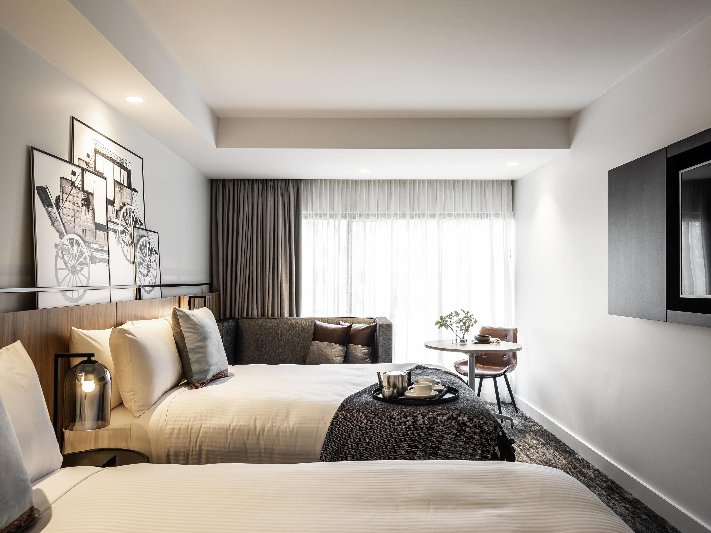 A hotel room with two beds, a desk, a chair, a TV, and artwork on the wall.