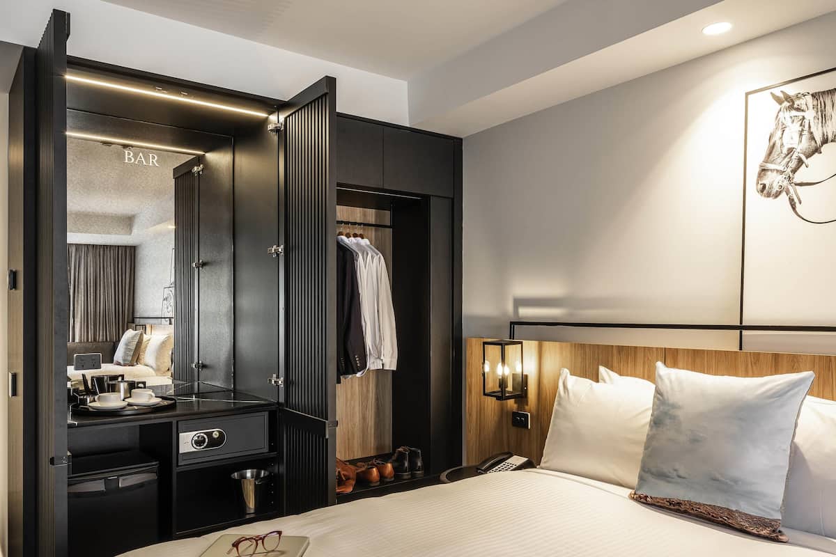 A hotel room with a bed, a desk with a laptop, a mirror, a wardrobe, and a bar area.