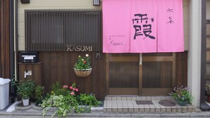 Una casa de huéspedes tradicional japonesa con una entrada de madera, un banderín rosa con caracteres japoneses y un pequeño jardín con plantas y flores en macetas.