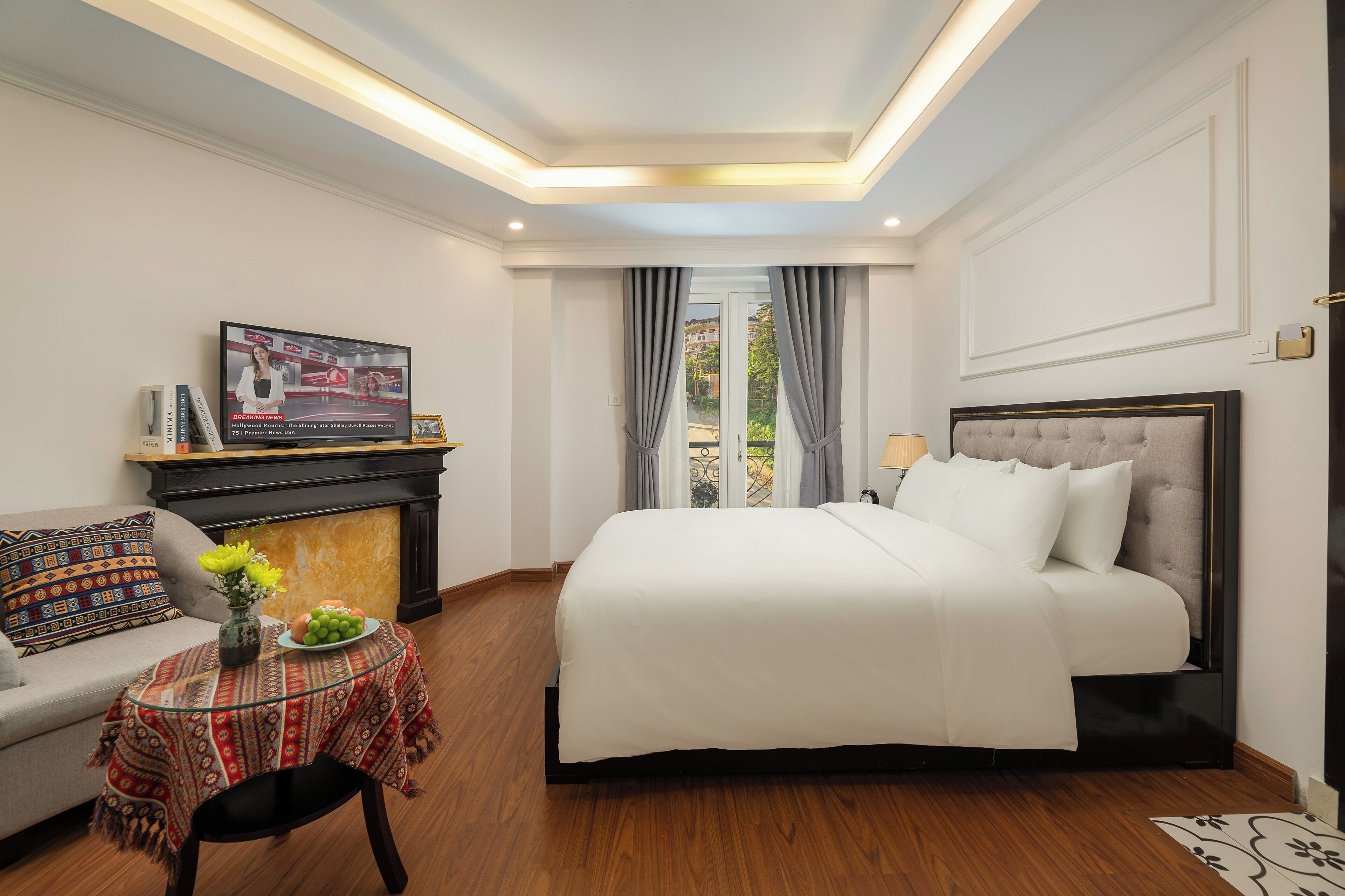 A hotel room with a large bed, a sofa, a small table with a vase and fruit, and a television on a stand.
