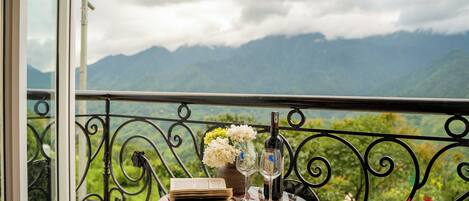 A balcony with a table set for two, featuring a book, a vase with flowers, and a bottle of wine.
