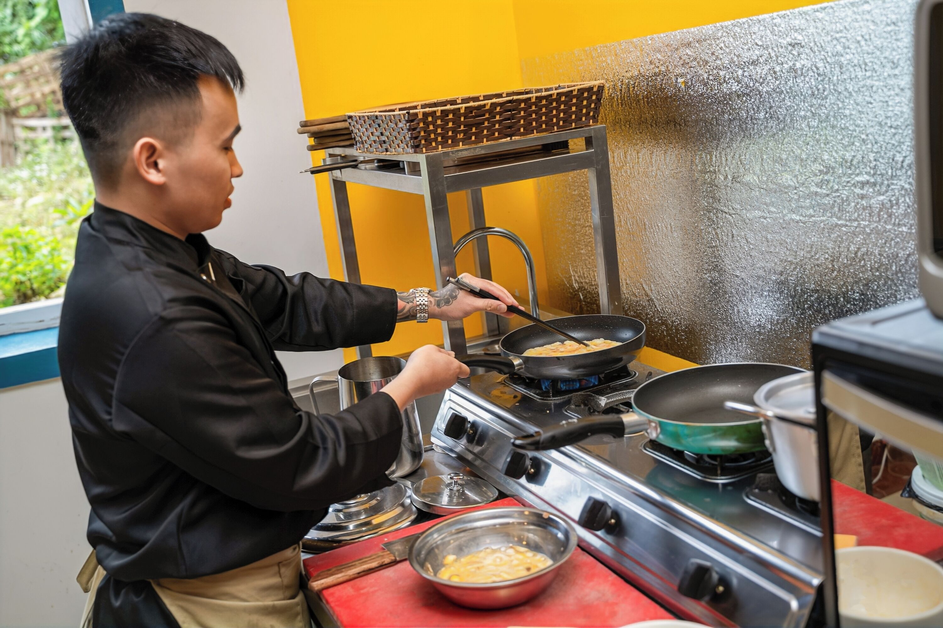 A person cooking on a stove with a basket on a metal stand and a window with greenery outside.