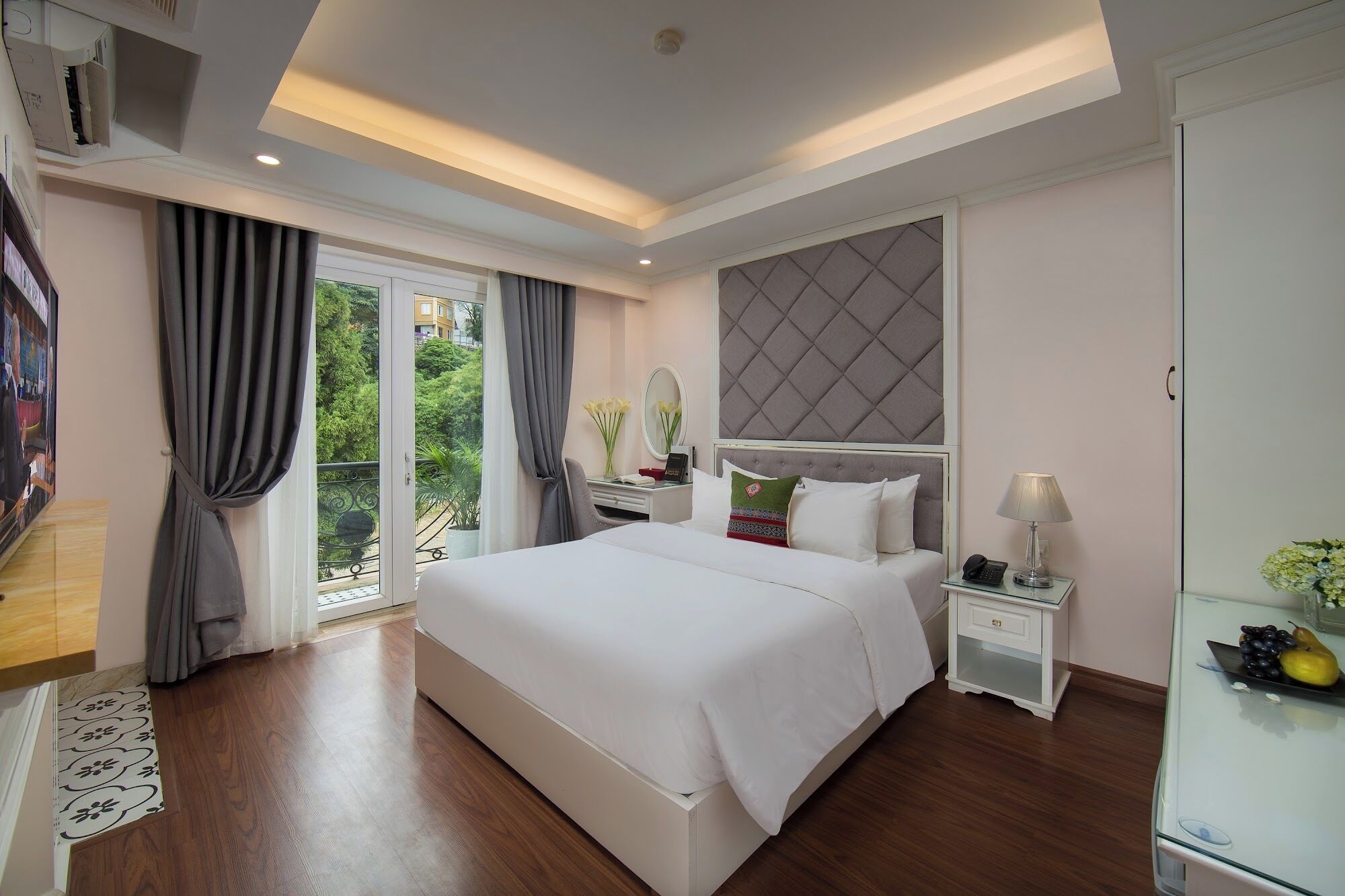 A modern hotel room with a large bed, a bedside table with a lamp, a mirror, and a view of greenery through the window.