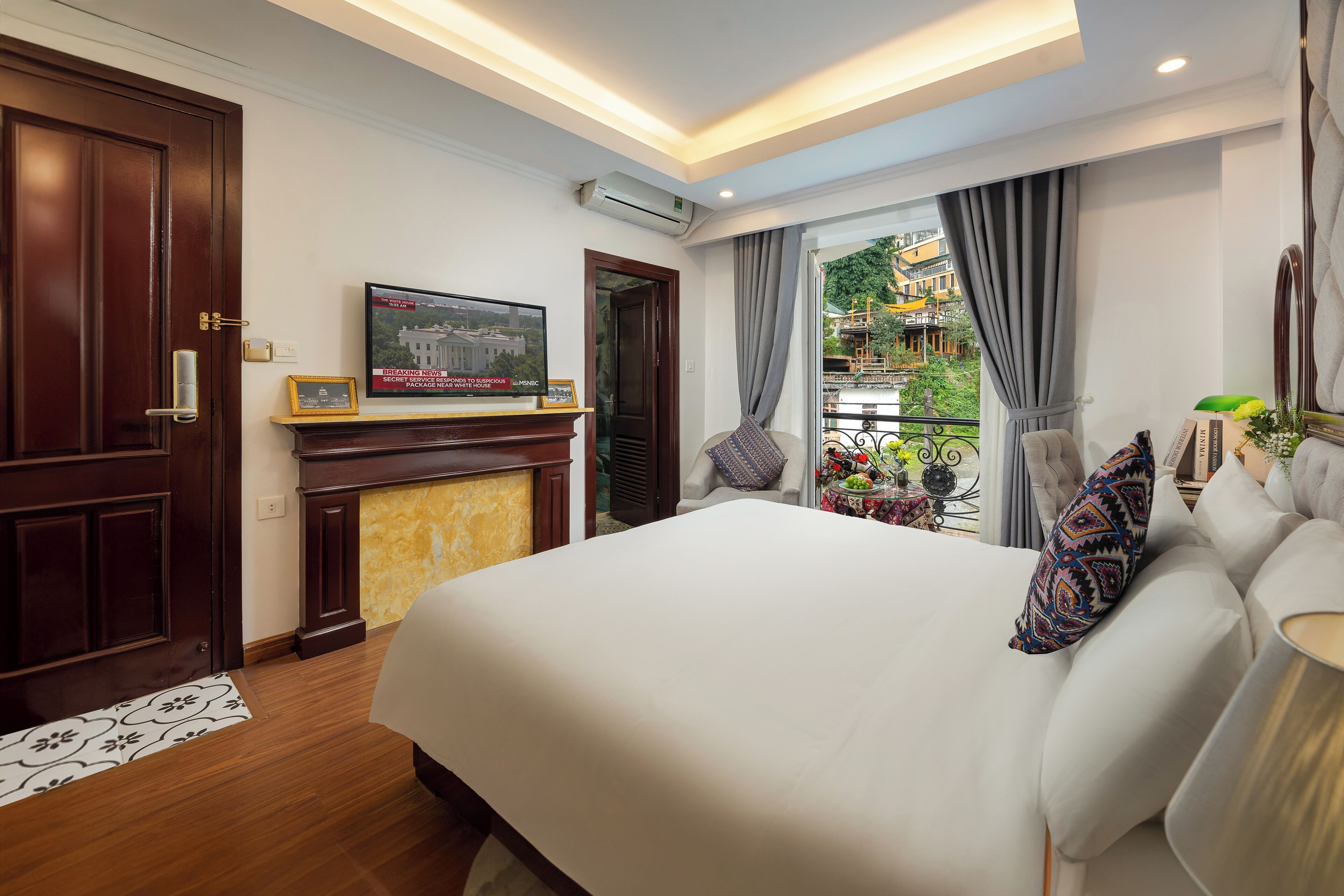 A hotel room with a large bed, a fireplace, a television, and a balcony with a view of a garden.