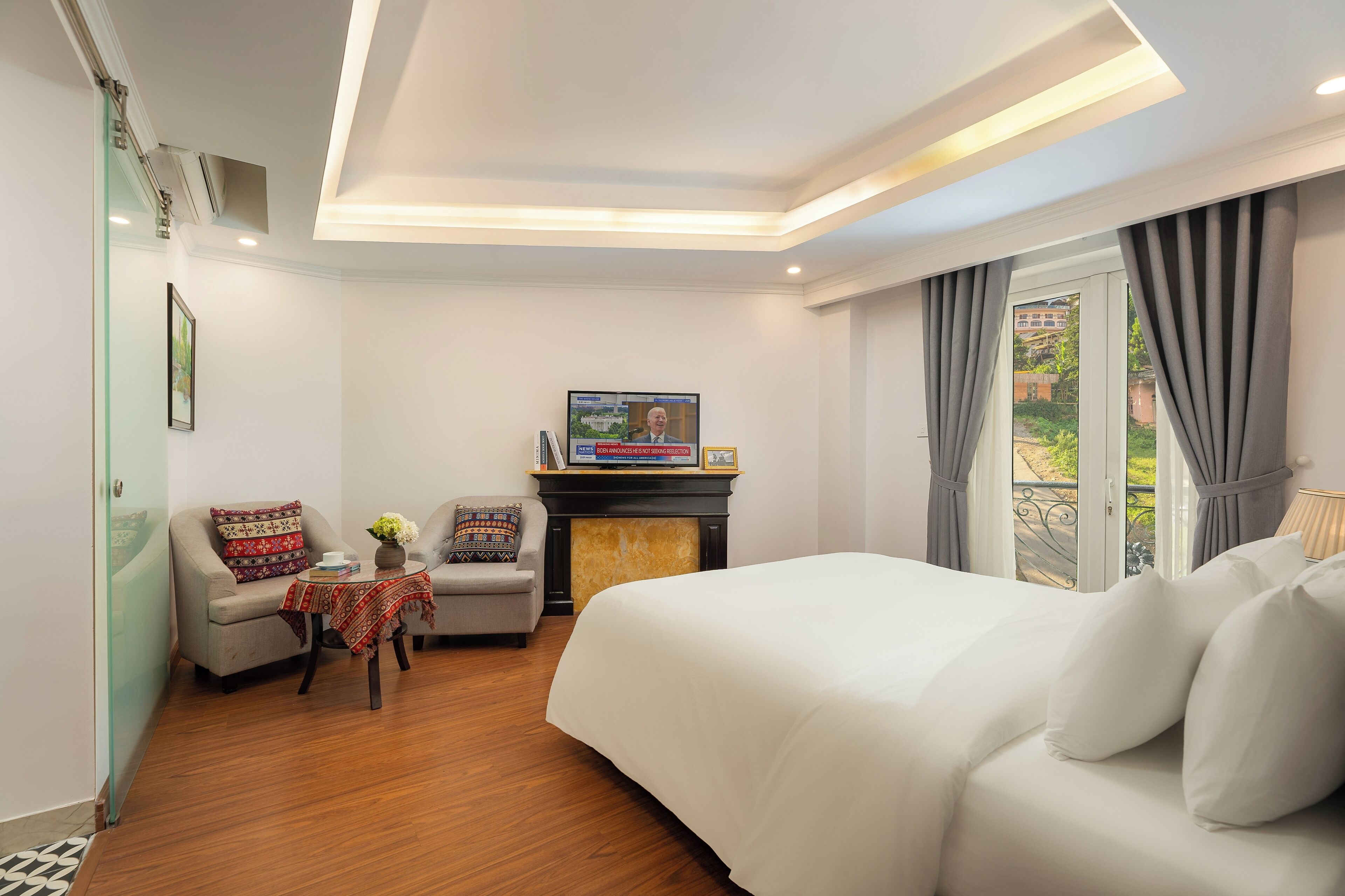 A hotel room with a bed, a sofa, a small table with a vase, a fireplace, and a television.