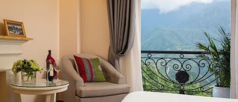 A room with a balcony, a chair, a table with a glass vase and wine bottle, and a view of mountains.