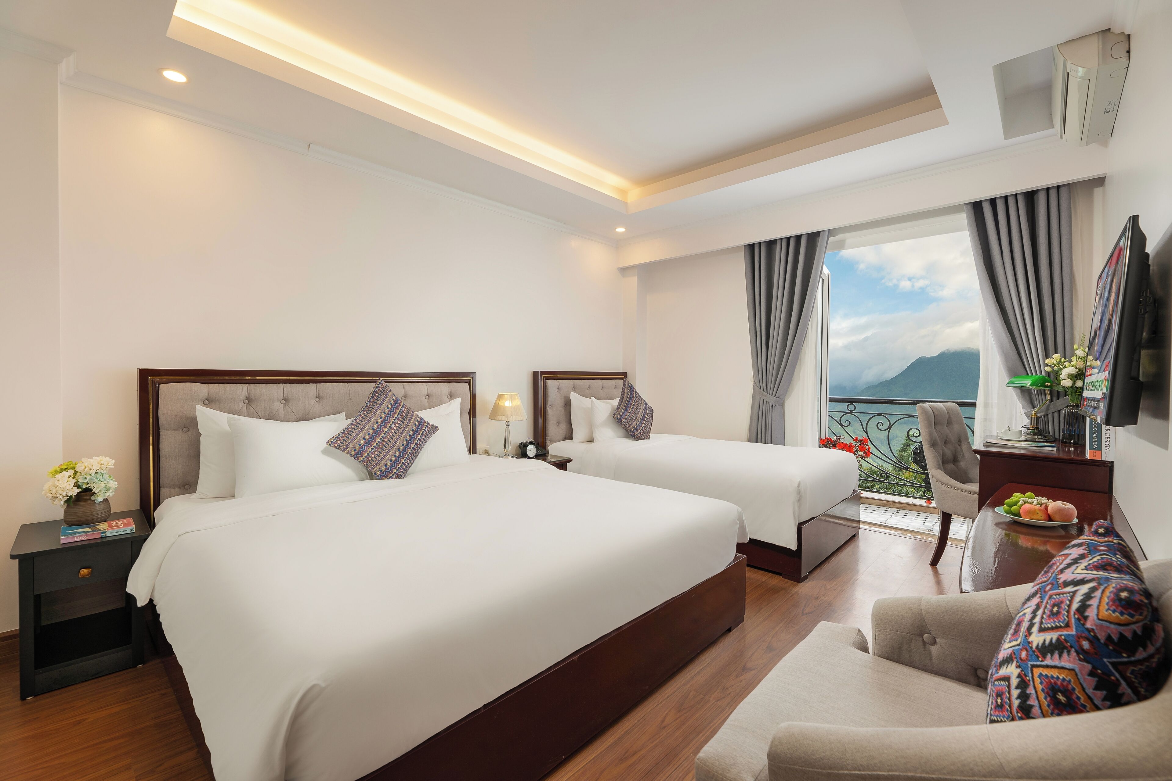 A hotel room with two beds, a sofa, a TV, and a balcony with a view of mountains.