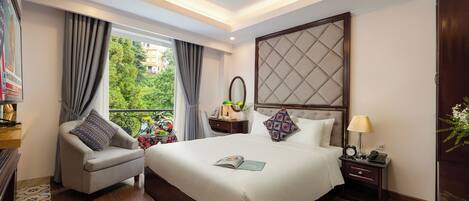 A hotel room with a large bed, a chair, a nightstand with a lamp, and a view of greenery outside.