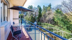 Ein Balkon mit Tisch und Stühlen, einem blauen Sonnenschirm und Blick auf Bäume und Grün.