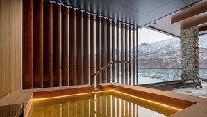 A modern spa area with a wooden bathtub, a stone wall, and a view of snowy mountains. - Yu Kiroro (Akaigawa)