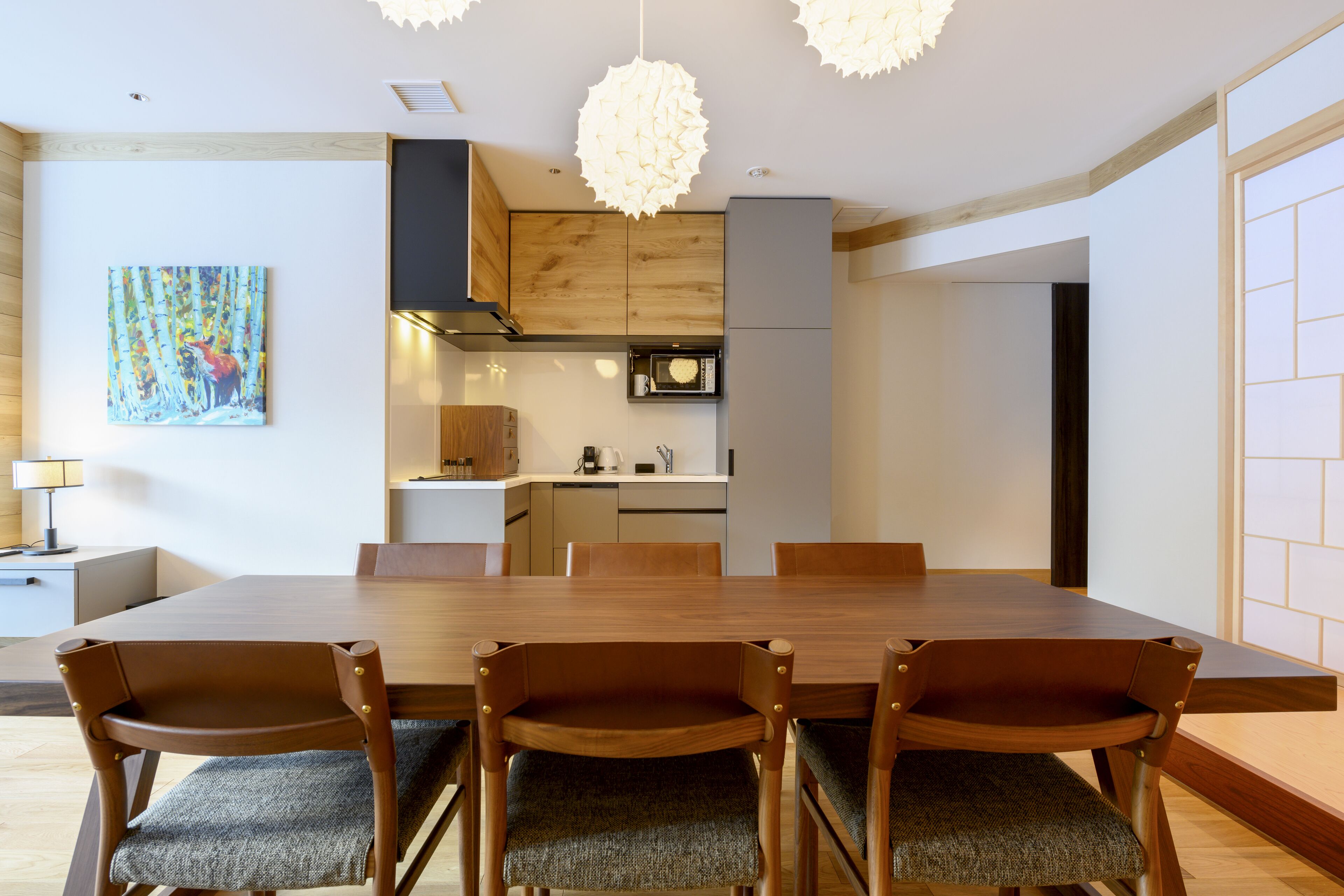 A modern dining area with a wooden table and chairs, a kitchen in the background, and a painting on the wall.