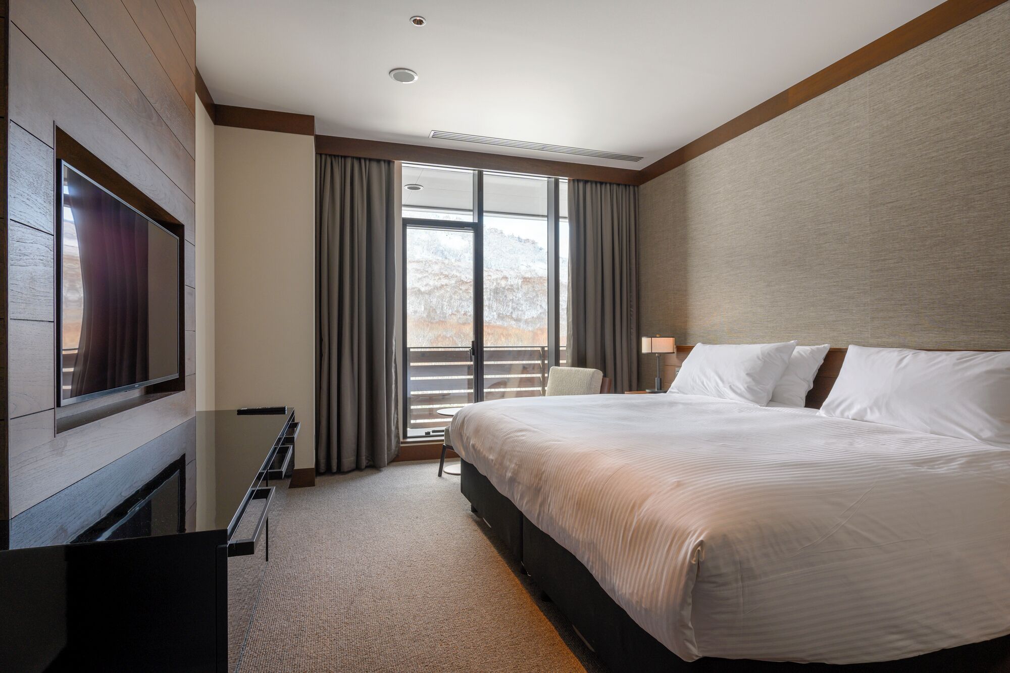 A hotel room with a large bed, a flat-screen TV mounted on the wall, a desk with a chair, and a view of a snowy landscape through the windows.