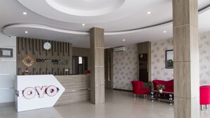 A hotel reception area with a modern design, featuring a reception desk with the hotel name, a clock, and a logo. There are red chairs and a small table with a plant.