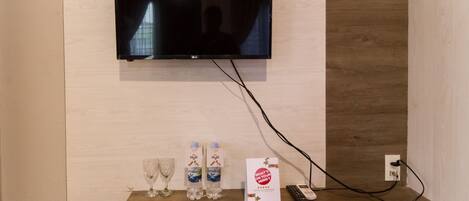 A room with a mounted flat-screen TV, two water bottles, a remote control, and a brochure on the floor.