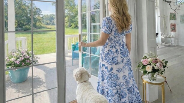 A woman in a blue floral dress opening a glass door to a patio with a dog, a potted plant, and a vase of flowers.
