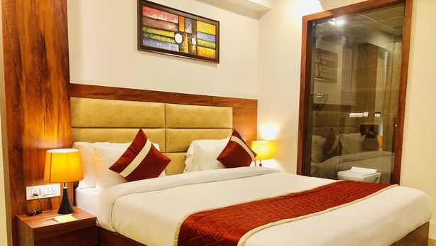 A hotel room with a large bed, wooden headboard, two bedside tables with lamps, a glass shower enclosure, and a colorful abstract painting on the wall. - Red Crown Hotel (New Delhi)