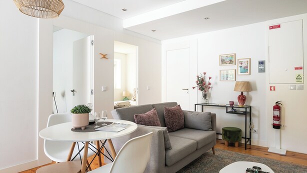 A modern living room with a grey sofa, white chairs, a small round table, and a coffee table. There is a plant on the table, a lamp on a side table, and a fire extinguisher on the wall.