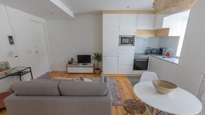 A modern apartment with a grey sofa, a small dining table with a bowl, a kitchen with white cabinets, a microwave, and a TV.