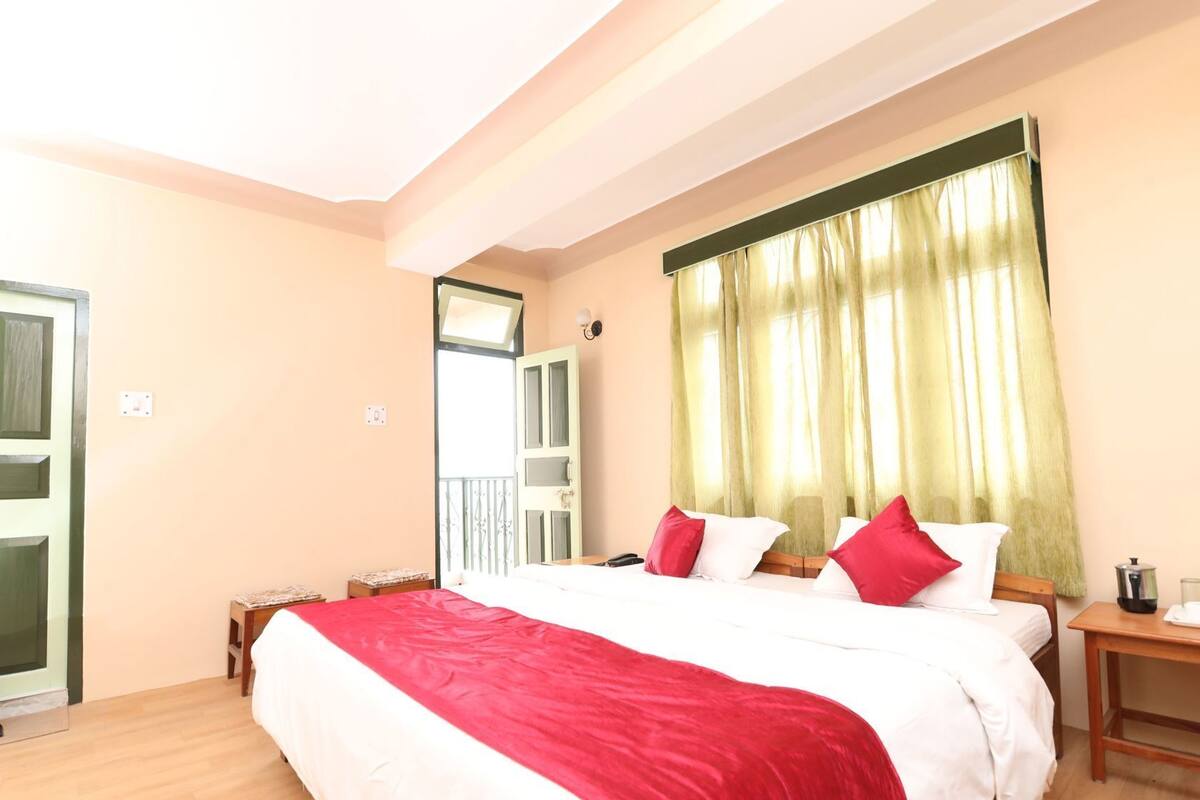 Habitación de hotel con una cama grande, una mesita de noche de madera, una pequeña mesa, una ventana con cortinas y una puerta.