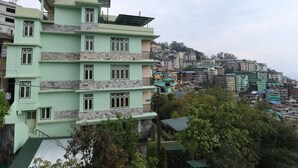A multi-story building with a green facade and stone accents, surrounded by densely packed hillside buildings. - Hotel Tenancy (Gangtok)