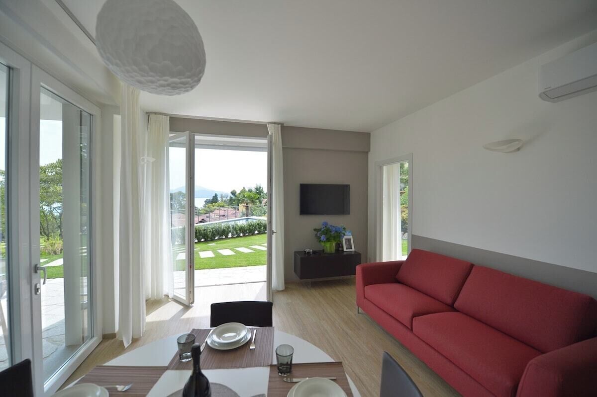 A modern living room with a red sofa, a dining table with chairs, a TV mounted on the wall, and a sliding glass door leading to a garden.