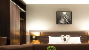 Una habitación de hotel moderna con un sofá marrón, una mesita con dos plantas en macetas, un cuadro en la pared y un cabecero con un arreglo de almohadas en forma de corazón.