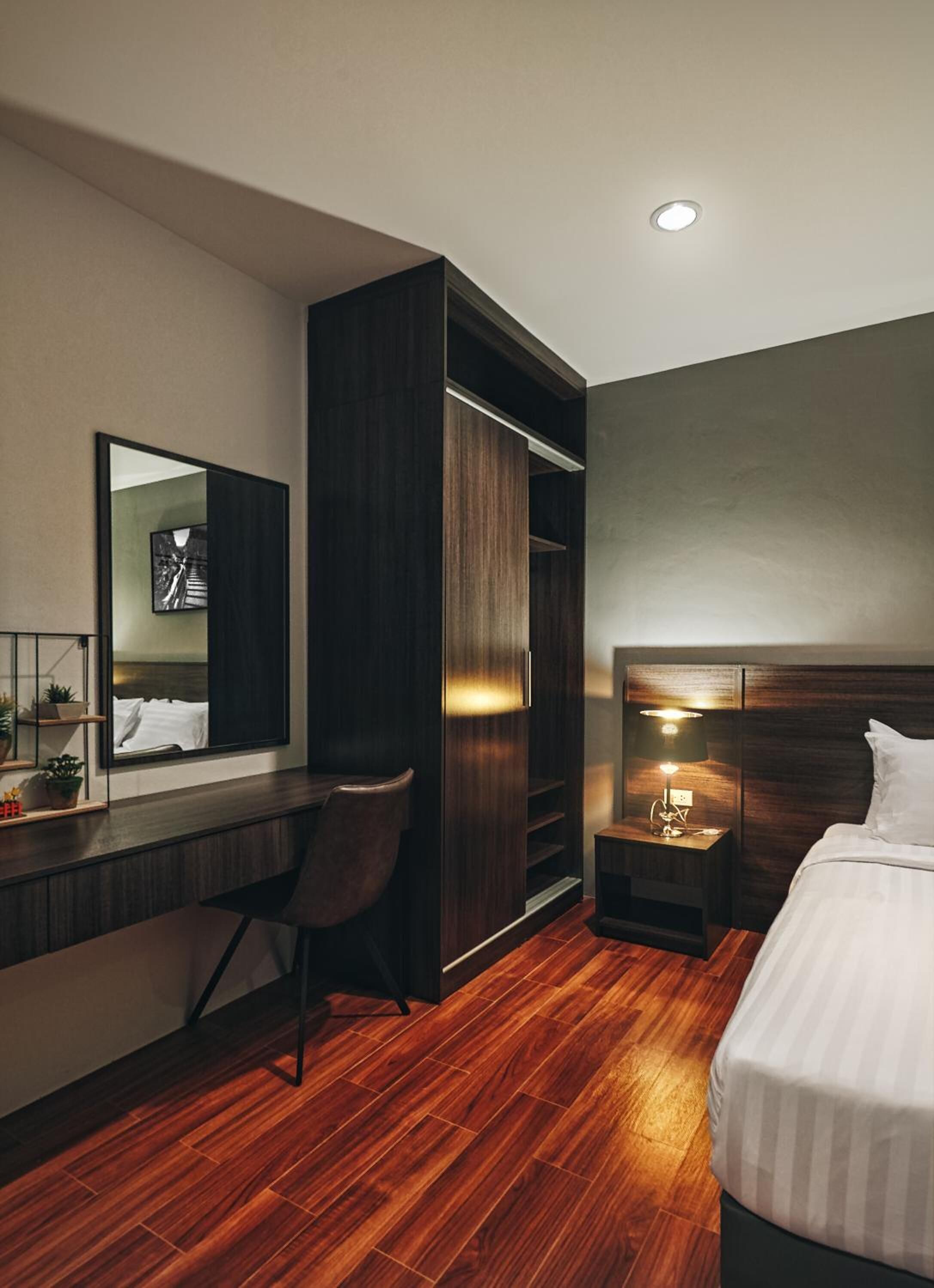A hotel room with a wooden floor, a bed, a desk with a chair, a wardrobe, a bedside table with a lamp, and a mirror on the wall.