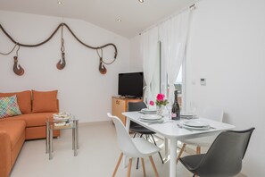 En moderne stue med sofa, sofabord i glas, spisebord dækket med tallerkener, og et fjernsyn i baggrunden.
