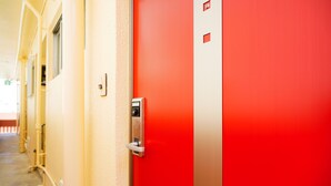 Una puerta roja con un picaporte plateado y una cerradura, parte de un edificio con fachada color crema y tuberías visibles.