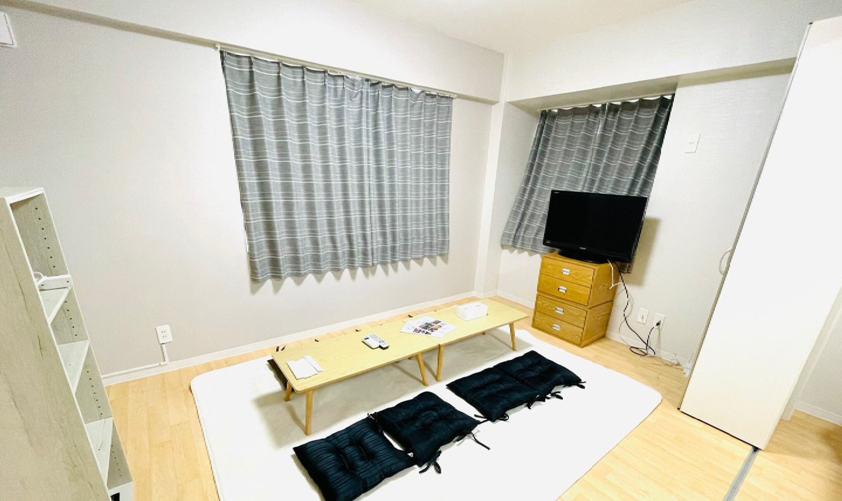 A small, sparsely furnished room with a wooden desk, two black cushions, a television on a wooden cabinet, and two windows with grey curtains.