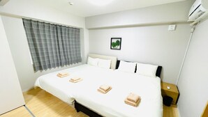 Una cama bien hecha con ropa de cama blanca y tres toallas dobladas, una mesita de noche con una lámpara, y una ventana con cortinas grises.