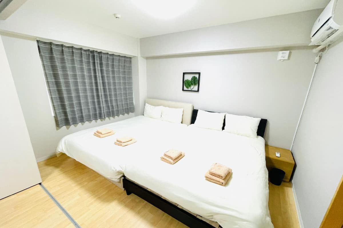 Uma cama bem arrumada com lençóis brancos e três toalhas dobradas, uma mesa de cabeceira com abajur e uma janela com cortinas cinzas.
