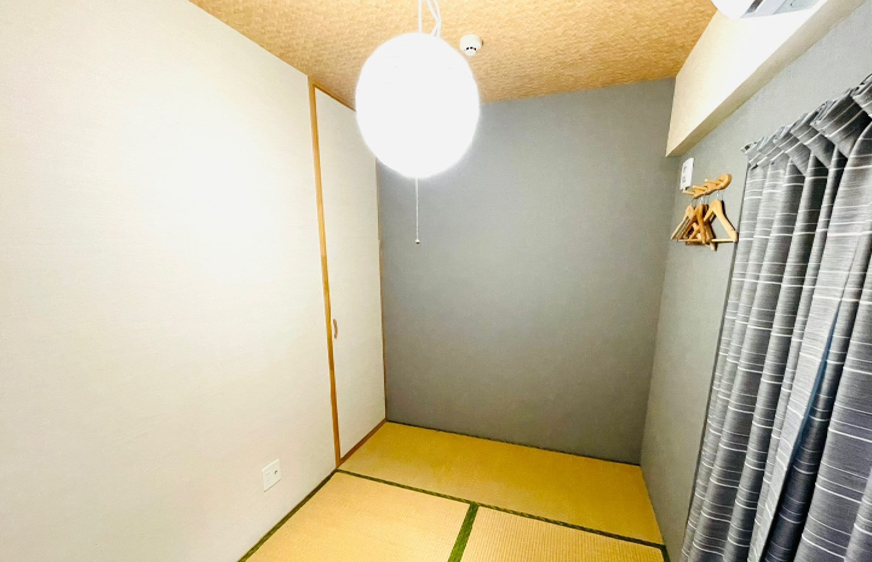 A small, sparsely furnished room with a tatami mat floor, a ceiling light, and a curtain on one side.