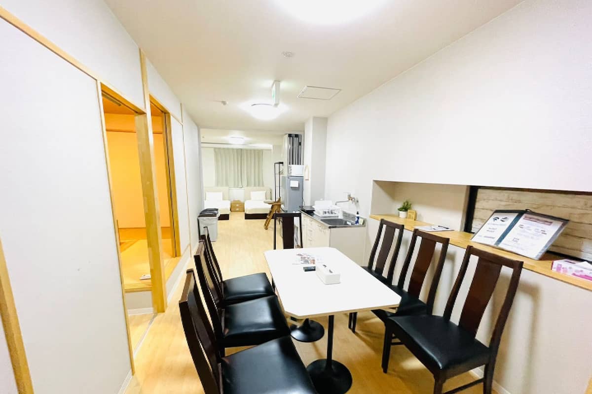 Quarto de hotel moderno com área de refeições, mesa branca e cadeiras pretas, cozinha compacta e uma pequena planta no balcão.
