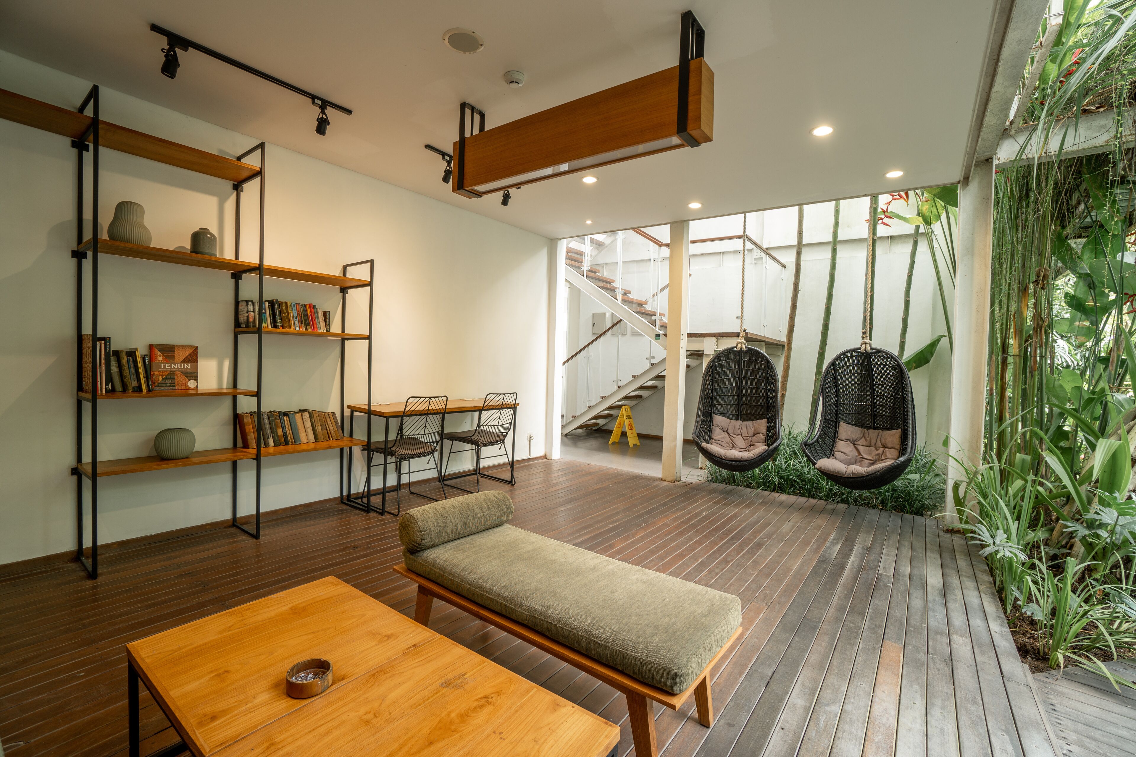 A modern interior with a wooden bench, bookshelves, a dining table, and a staircase leading to a balcony with hanging chairs.