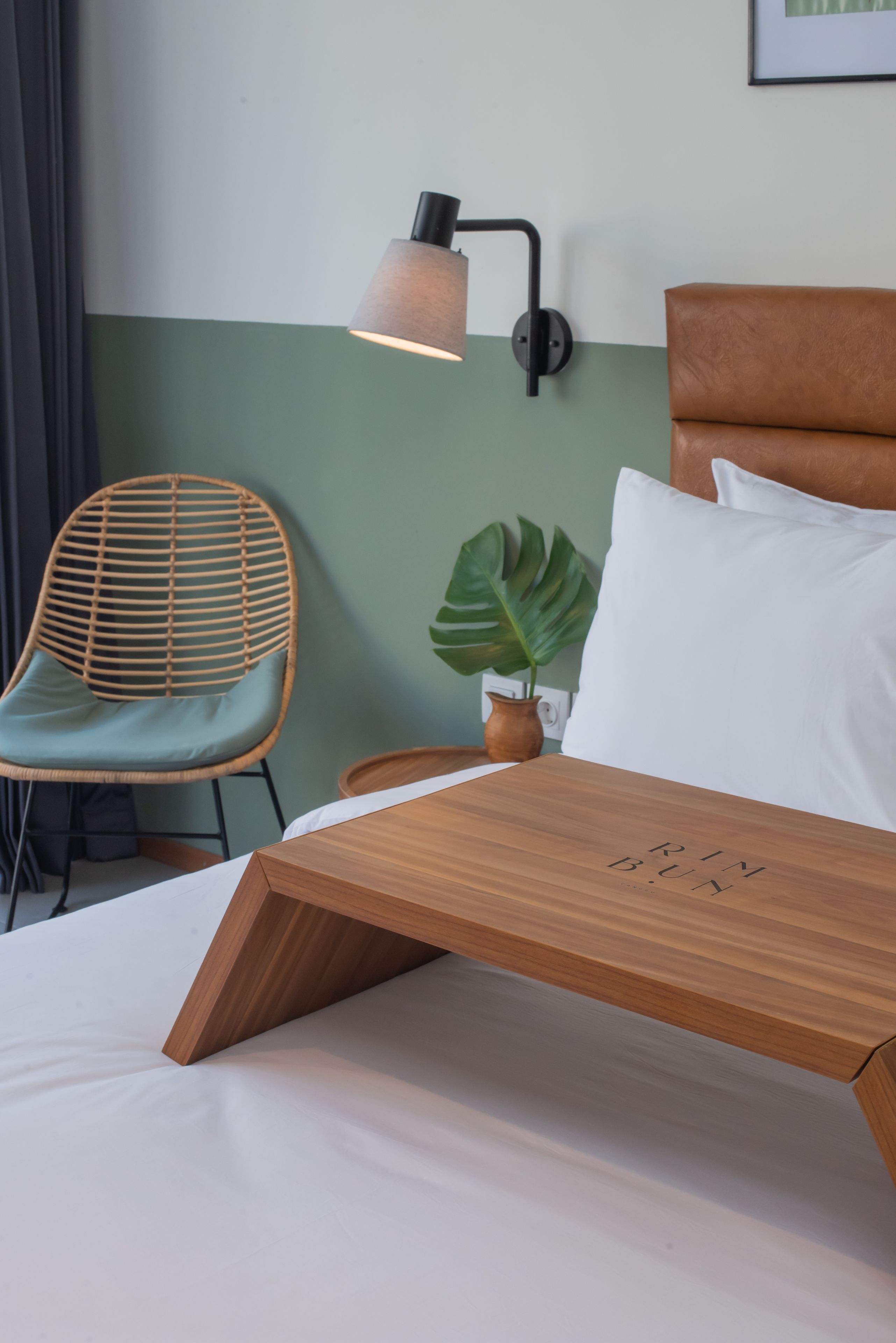 Une chambre d’hôtel bien rangée, avec un lit en bois, une chaise en osier, une lampe fixée au mur et une plante en pot.