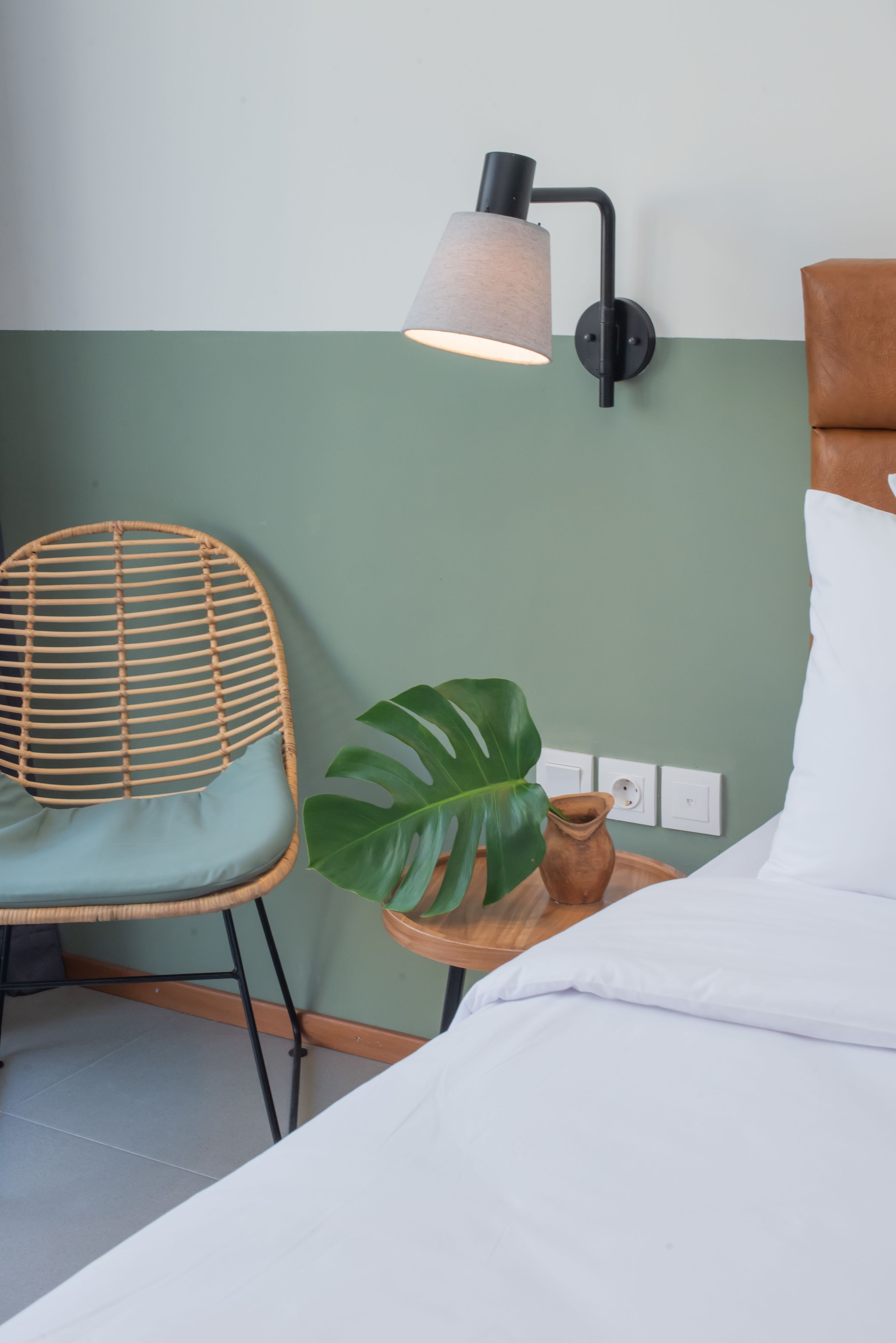 A room with a bed, a wicker chair, a potted plant, and a wall-mounted lamp.