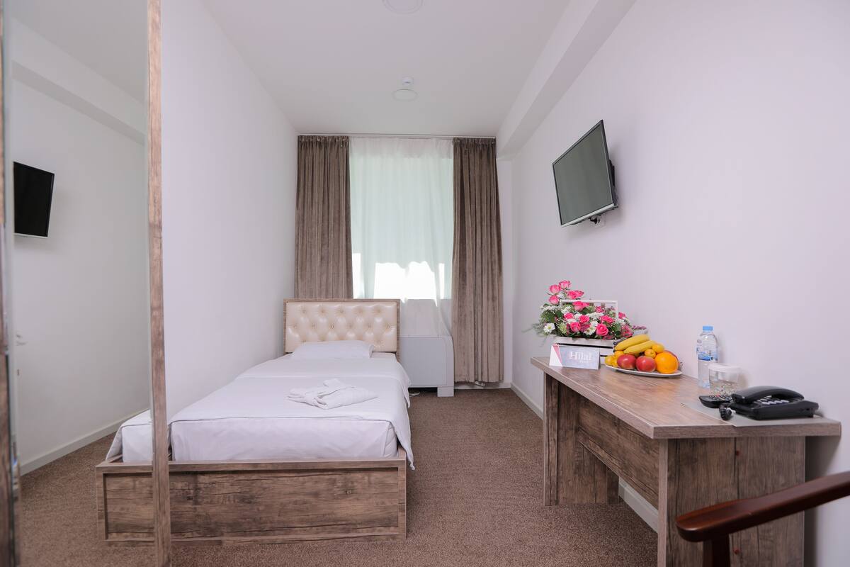 A hotel room with a single bed, a wooden desk with a telephone and a fruit bowl, and a TV mounted on the wall.